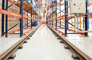 Trikryptis automatinis stelažų krautuvas padėklams važiuoja tarpueiliu dviem apatiniais bėgeliais